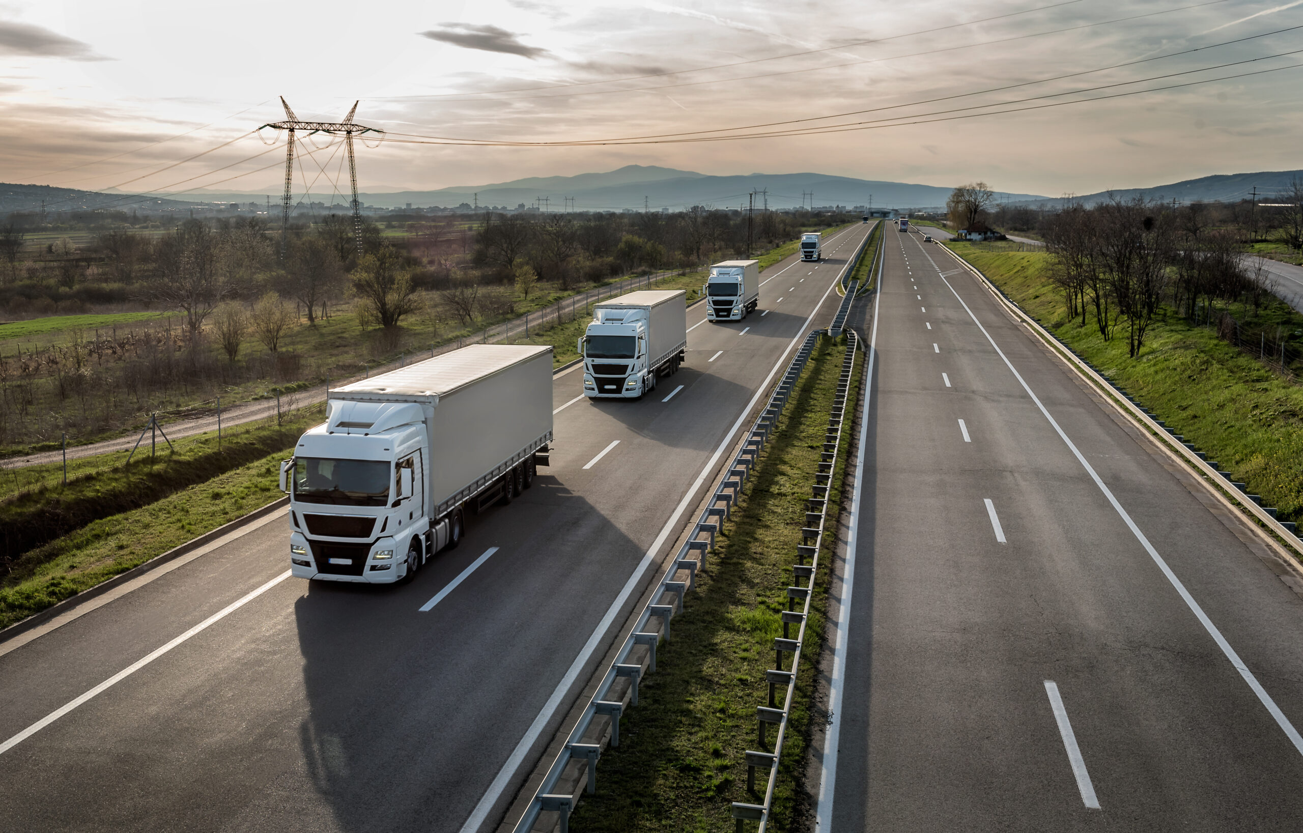Transport de marchandises : trois camions blancs sur une autoroute moderne, avec pylônes électriques et collines au loin.